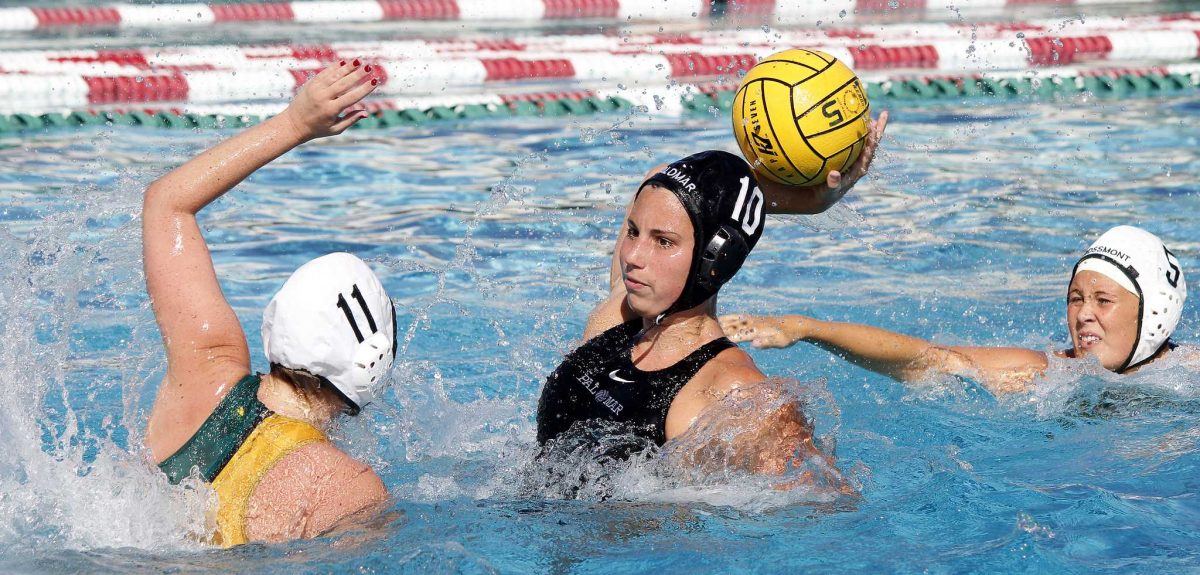 A female Palomar water polo player tries to throw the ball in her right hand as two opponents try to block her from the left and right.