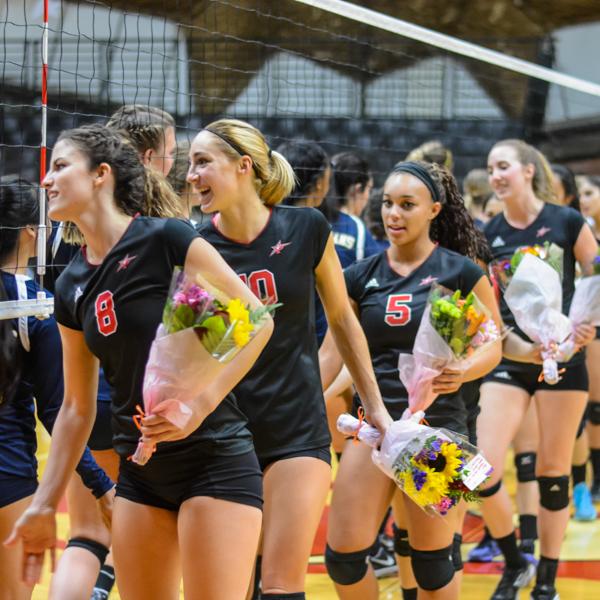 Palomar Women's Volleyball team line in a queue and walking by the net to give low-fives to the opposing team. Some of them carry a flower bouquet.