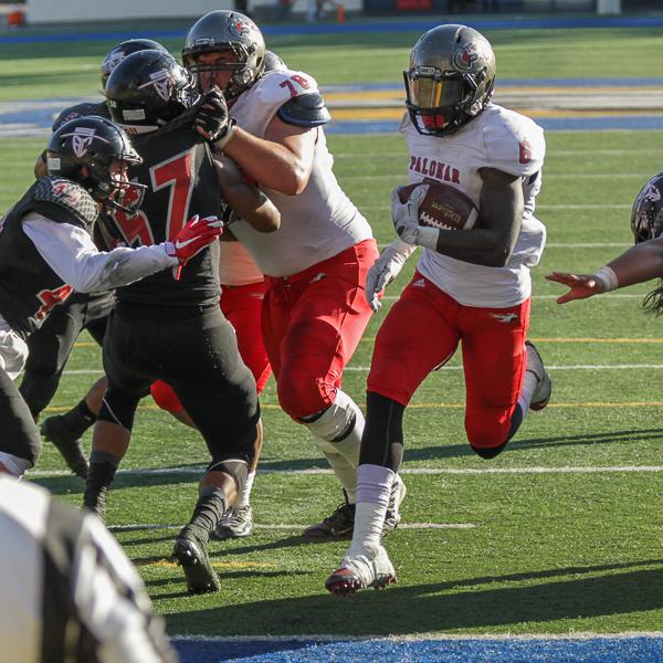 A Palomar football player runs with a football tucked in his left arm. Several players tackle each other on the sides.