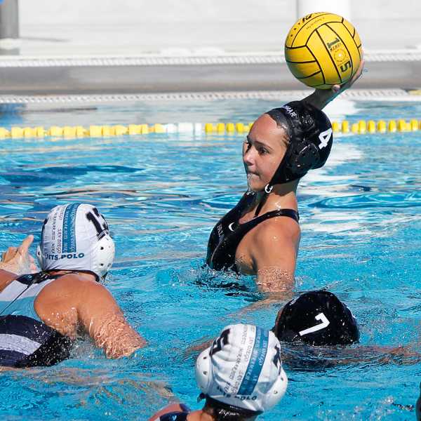 A female Palomar water polo player tries to throw the ball in her right hand as an opponent tries to block her from the left.