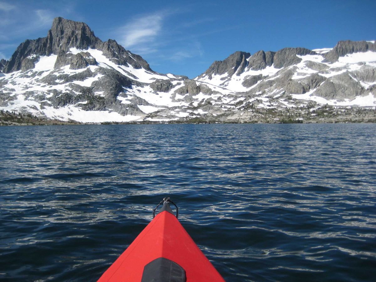 The tip of a red canoe peeks at the bottom of the photo, which is floated on a large body of water with a cobalt mountain range in front of it, coated here and there with snow.