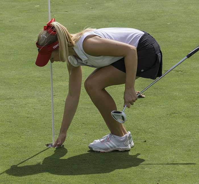 Palomar golfer Brittany Bilek picks up a golf ball from a hole on a golf course. She wears a red cap, white top, and black shorts.