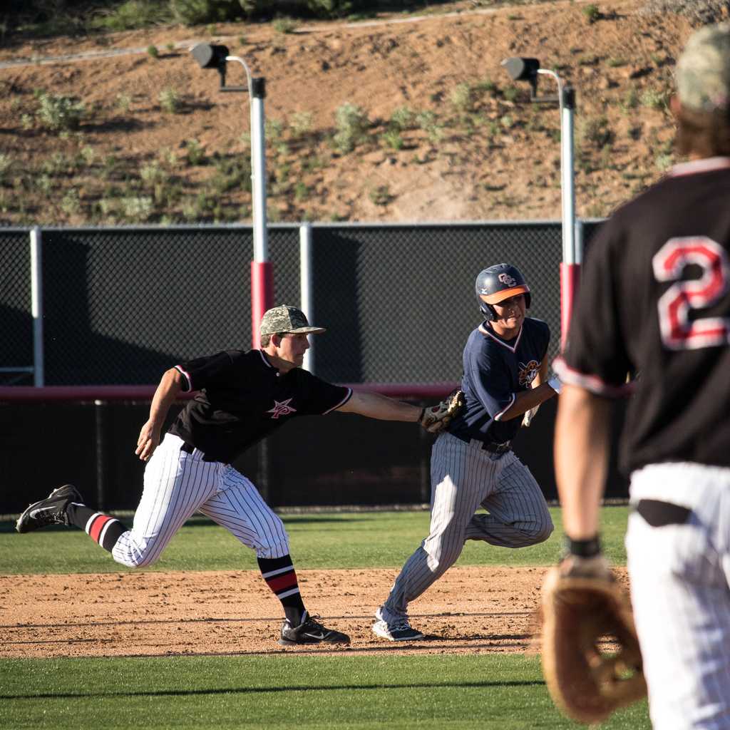 A male Palomar baseball player tags an opposing player as he runs after him. A teammate stands in the foreground (blurred), watching them.