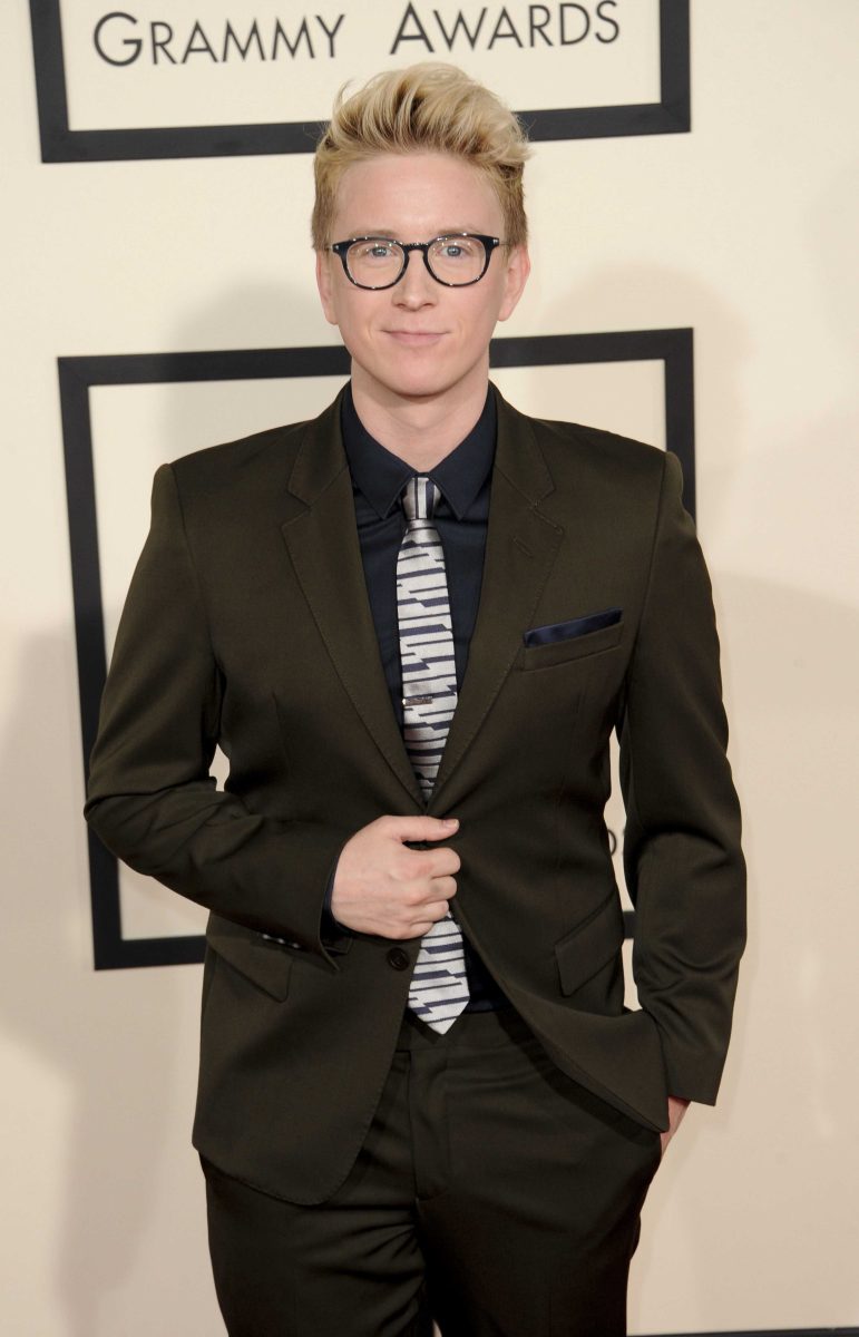 A man in a brown suit and a piano-key patterned tie stands with his left hand in his pants pocket and his right hand clasping on his suit at his abdomen.