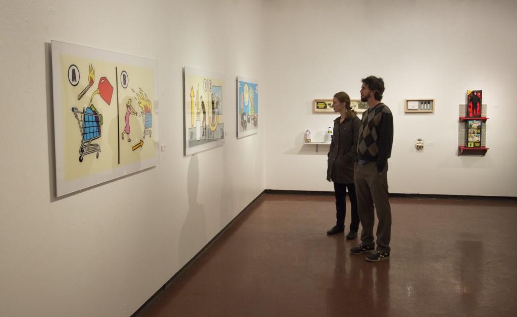 A woman and a man stand next to each other and look at three drawings on a wall.