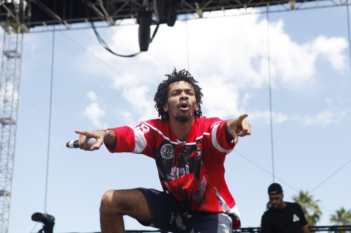 A man in a red shit points with both hands to his audience with his mouth in an O shape. A DJ is in the background.