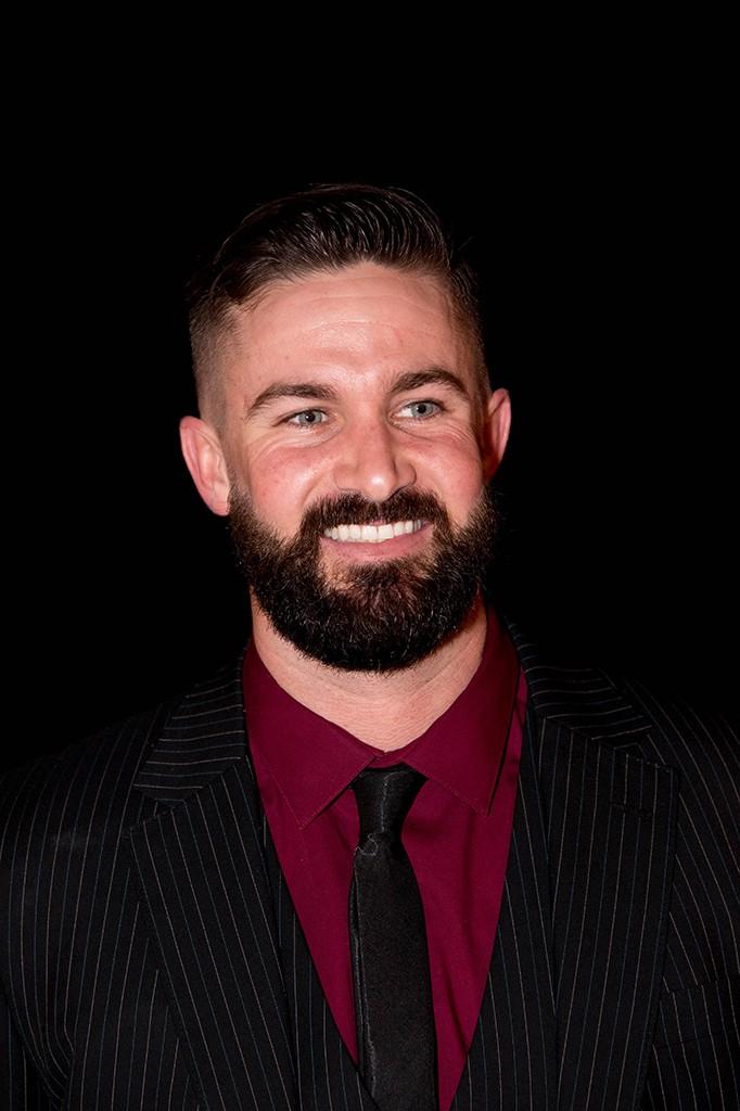 Palomar College inducted former Baseball player Nick Vincent smiles, wearing a black suit with thin, vertical dark yellow stripes, a red shirt, and a black tie.