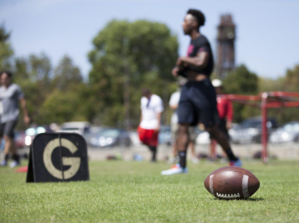 A football lies on the grassy ground in the lower right as a Palomar football player in plain clothes walk a few feet behind it (blurry).