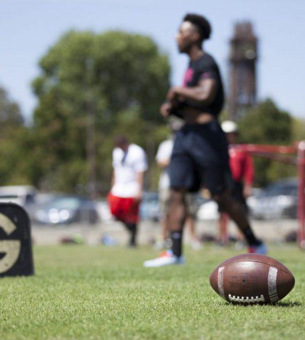 A football lies on the grassy ground in the lower right as a Palomar football player in plain clothes walk a few feet behind it (blurry).