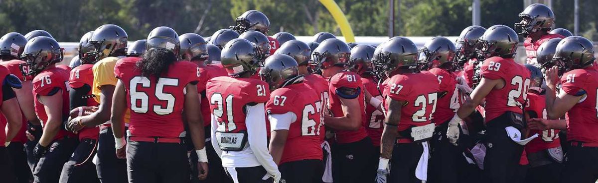 A team of Palomar football players gather on a field, wearing red jerseys, black game pants, and gray helmets.