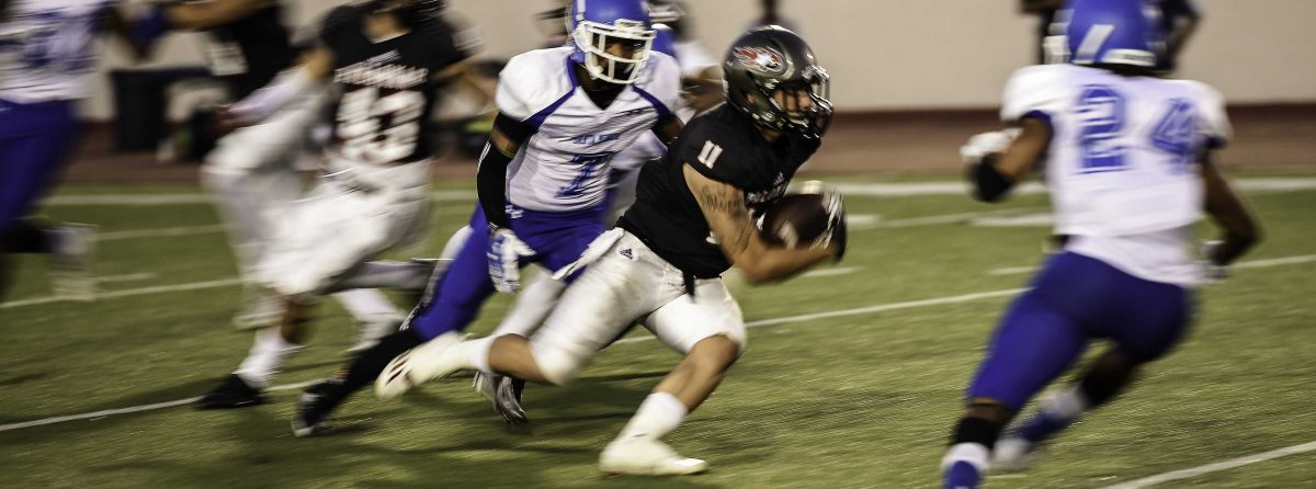 A Palomar football player dashes between two opposing players with a football in his right arm (blurry shot).