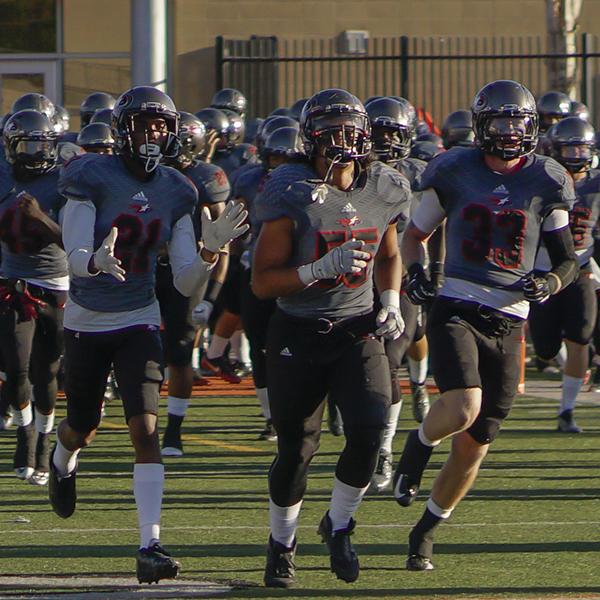 A team of Palomar football players runs out to a football field, wearing gray jerseys, black game pants, and gray helmets.