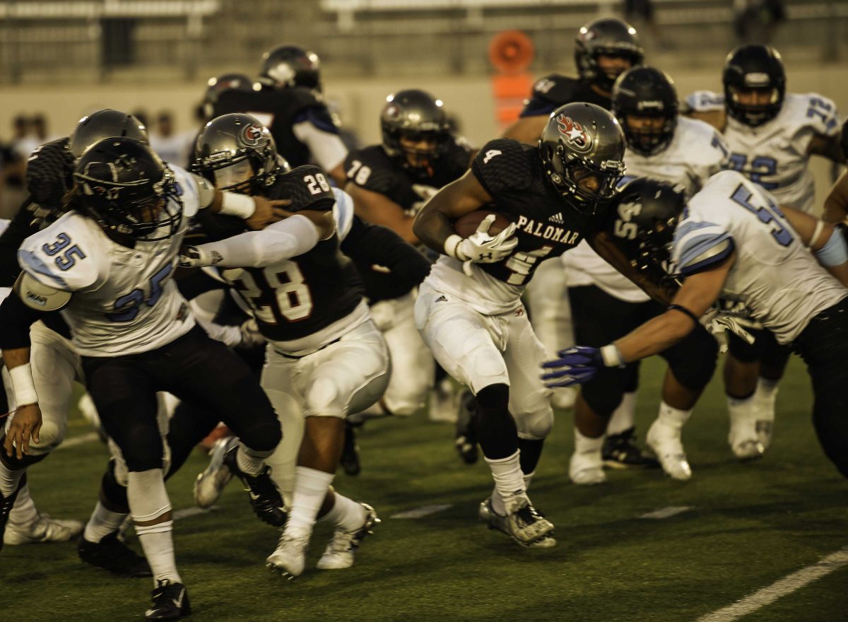 A Palomar football player runs with a football tucked in his right arm amidst a group of players tackling each other.