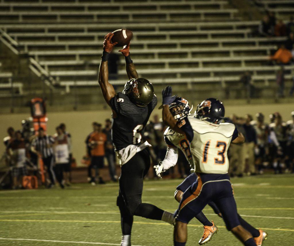 A Palomar football player catches a football with both hands above him as two opposing players tries to tackle him from behind.