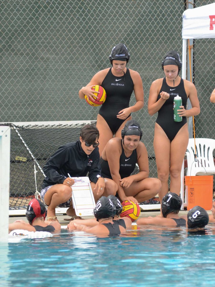 Palomar Head Coach Jackie Puccino crouches by a swimming pool, showing her water polo players strategies with a paper on a clipboard.