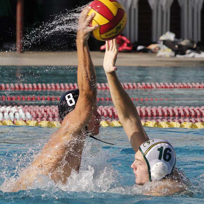 A male Palomar water polo player throws a ball with his right hand as an opponent tries to block it with his right hand.