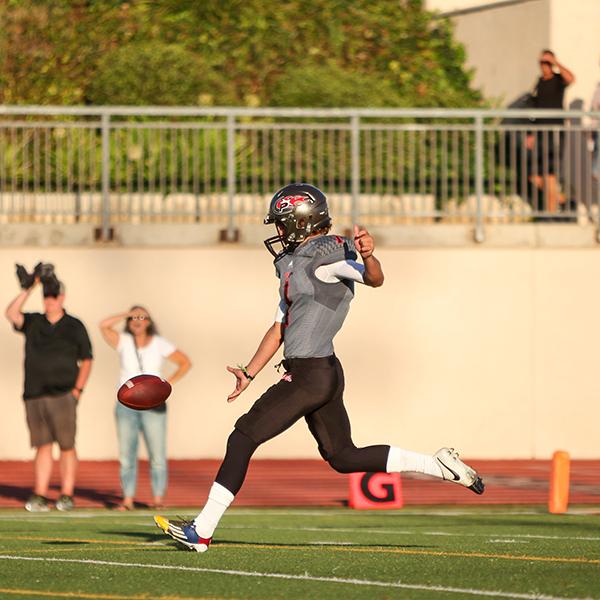 A Palomar football player drops a football in front of him and brings his right leg back to kick. Several people stand and watch in the background.