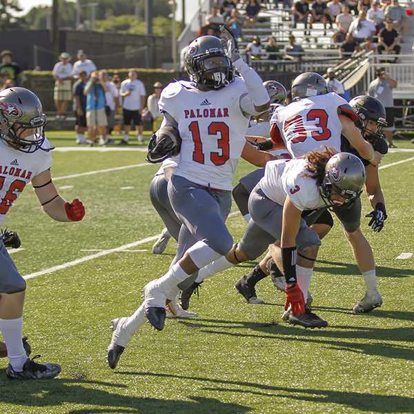 A Palomar football player hops up as he carries a football in his right arm. Several other player tackle each other in the background.