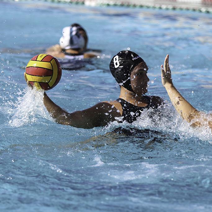 A female Palomar water polo player throws a yellow ball with her right hand in a pool as an opponent tries to block her on the right.