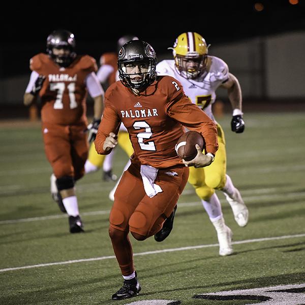 A Palomar football player, wearing an entire red uniform, runs and carries a football in his left arm. An opposing player and two team players run behind him.