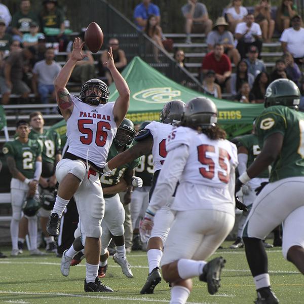 A Palomar football player catches a football with both hands with his right knee up while several players from both teams run toward him front his front and behind. A crowd of people watch from the metal bleachers in the background.