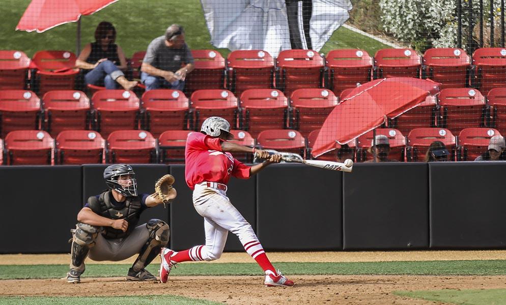 A male Palomar baseball player in a red jersey and white pants swings a baseball bat with the baseball inches from the bat. A catching squats behind him, and two people sit on the red seats in the background.