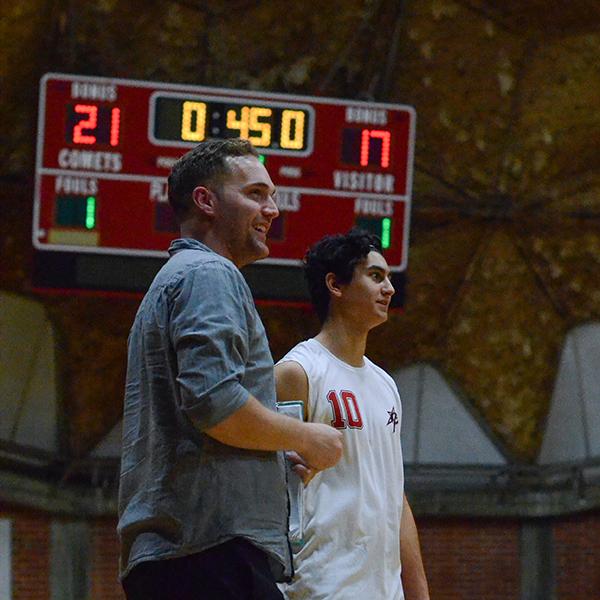 Palomar's Men's Volleyball Head Coach Bryan Campbells stands next to a malel volleyball player at The Dome. A scoreboard is in the background.