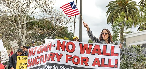 A woman holds a large horizontal sign that says, "NO BAN NO WALL SANCTUARY FOR ALL" and holds an American flag with the right hand.