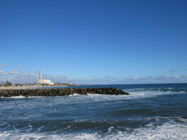 Ocean waves hit a rocky ledge on a sunny day. A factory with a single chimney is in the far background.