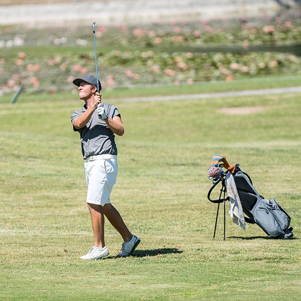 Palomar's Mitch Cannon swings a golf club on a golf course. A golf bag filled with colorful clubs is behind him.