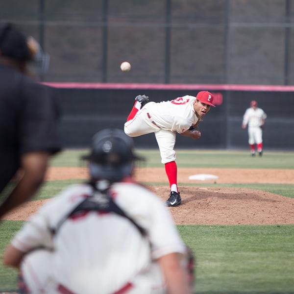 A male Palomar baseball player pitches a baseball toward the catcher and umpire in the foreground (blurry). His right leg is kicked out behind him.