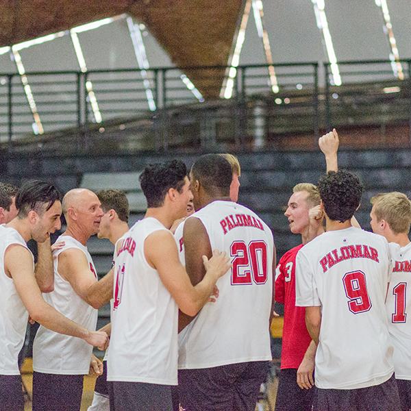 A group of male Palomar volleyball players gather around. They all wear white jerseys except for one guy to the right who is wearing a read jersey.