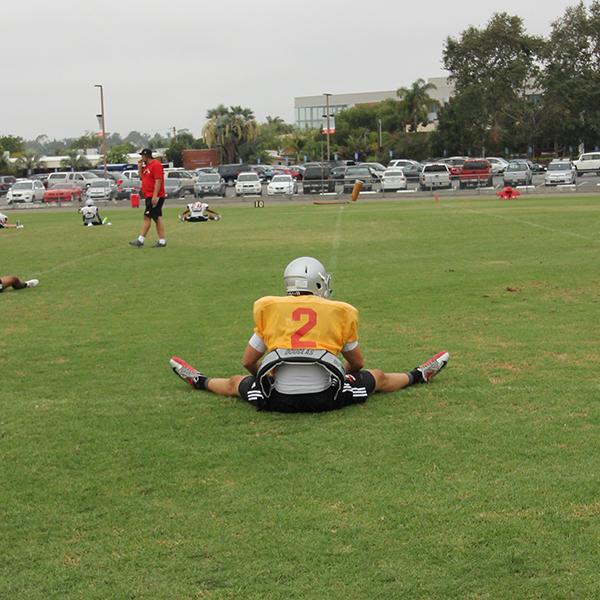 A Palomar football player in an orange-yellow jersey and black shorts sits on the field with his legs stretched out to his sides like a V shape.