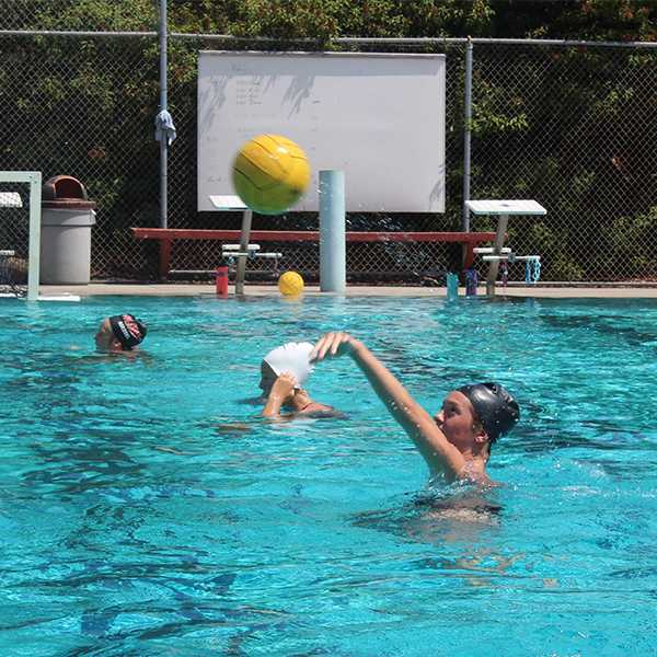 A female Palomar water polo player throws a ball with her left hand. Two other players are in the background in the pool.