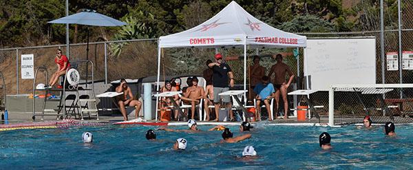 Several water polo players are in the pool as other players and a coach are sitting and standing beneath a white tent at the edge of the pool.