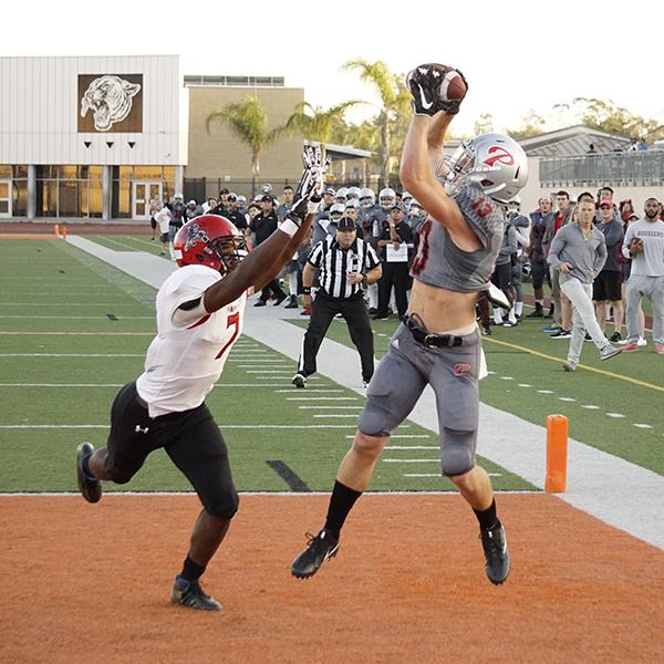 A Palomar football player jumps up and catches a football with both hands as an opposing player tries to intercept with his both hands. A referee and a crowd of Palomar players and coaches stand in the background and watch them.