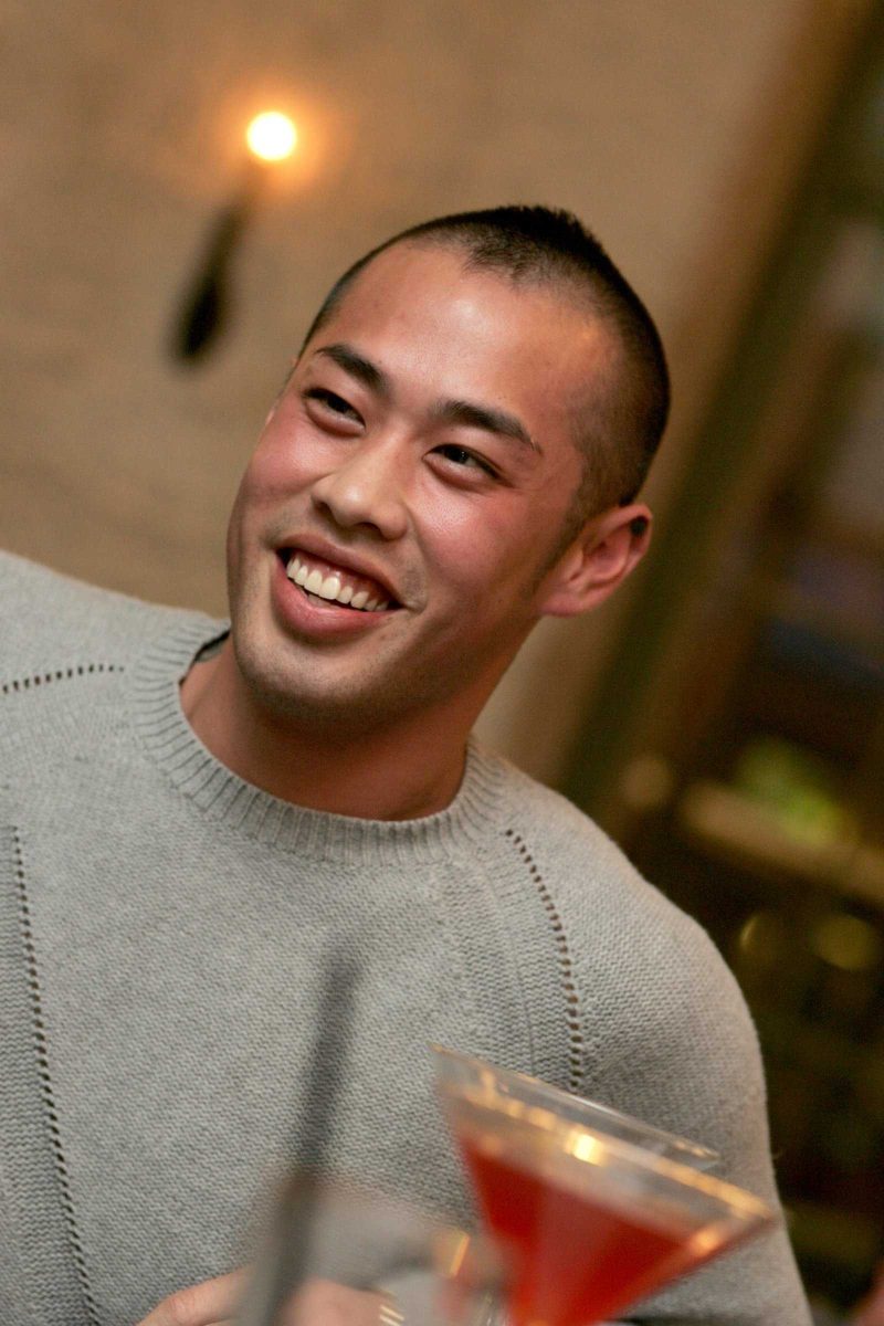 A man smiles at a cafe with a glass of red drink in front of him.