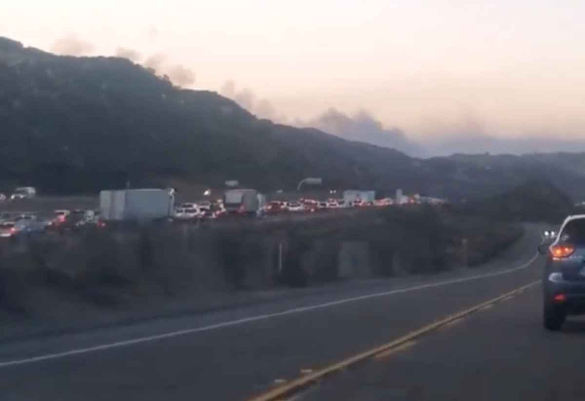 A traffic jam on a highway in the distance with a single-lane country road in the foreground that winds to the right. The rear of a minivan is half-shown at the right. A small mountain range is in the background with fire smoke behind it.