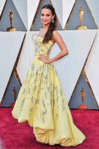 Actress Alicia Vikander stands on a red carpet and wears a long yellow dress with some text patterns and her left hand on her hip.