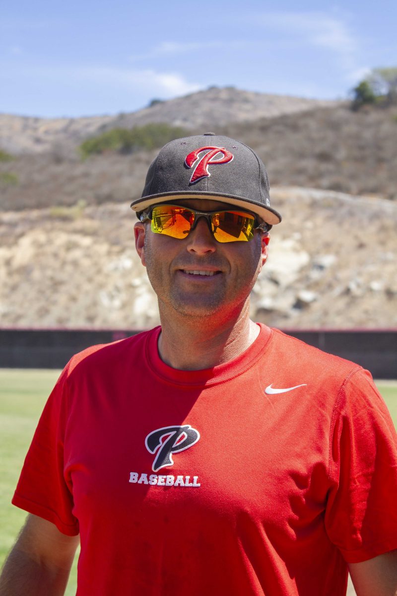 Palomar baseball coach Ben Adams stands on a baseball field, wearing a red T-shirt with the Palomar Baseball logo at the front and a Nike logo on the upper left chest, a black cap, and dark yellow sunglasses.