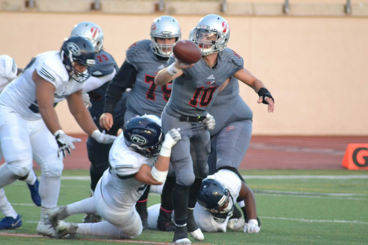 A Palomar football player gets tackled by an opposing player as he runs and carries a football in his right hand. A group of player runs behind them.