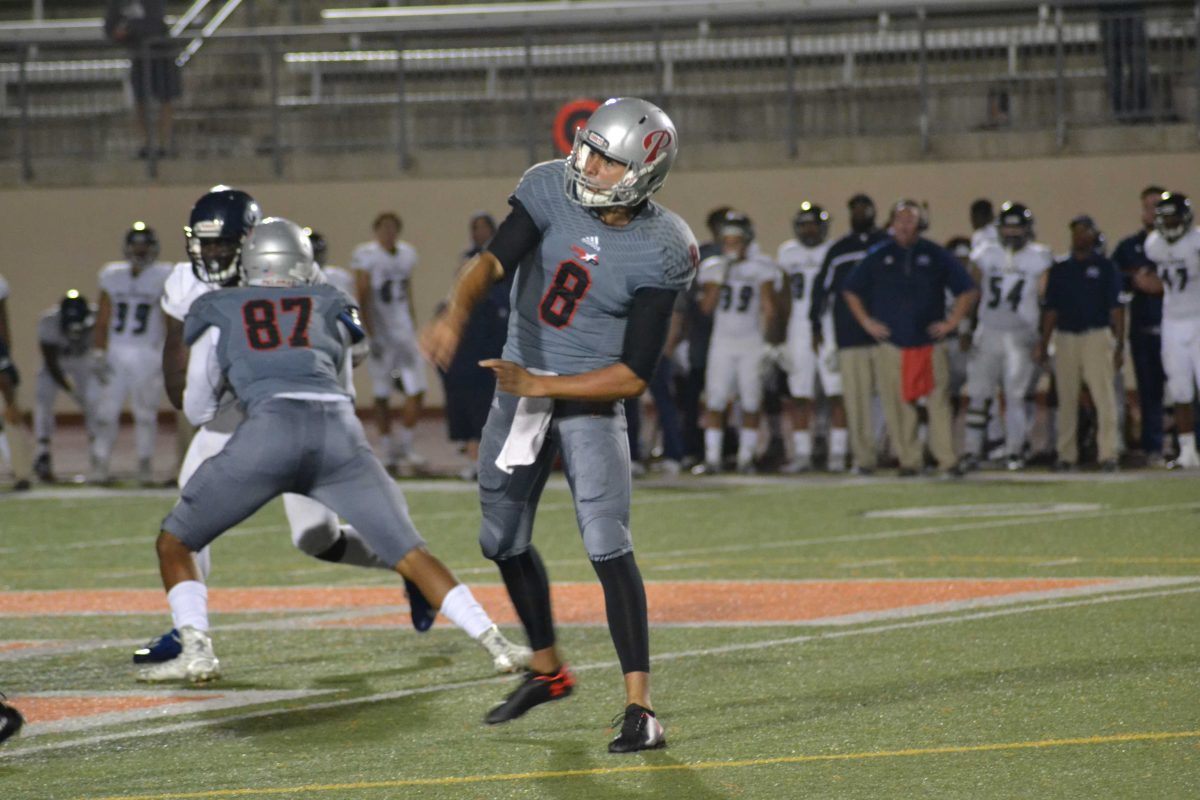 A Palomar football player throws a football with his right hand as two players tackle each other in the background to his right. A group of players, referees, and coaches stand in the background.