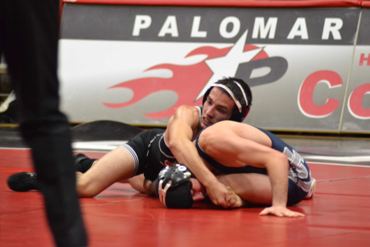 A male Palomar wrestler pins an opponent on the ground by wrapping his arms around his body while his opponent seems to be leaning forward and sitting cross-legged. A referee's leg in on the left.