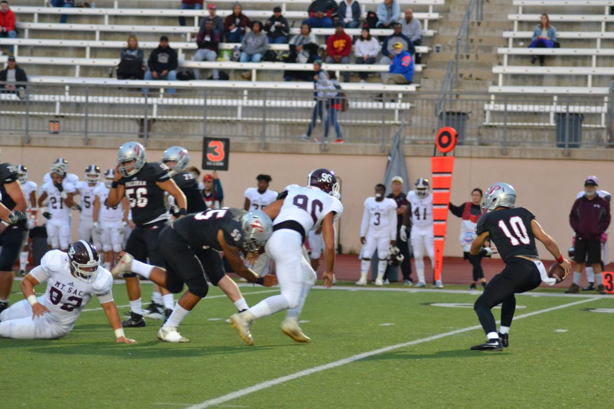 A Palomar football player on the right tries to dodge an opposing player who is running toward him. Another Palomar player tries to tackle that opponent from behind as other players tackles each other behind him. A small crowd sits on bleachers in the background.