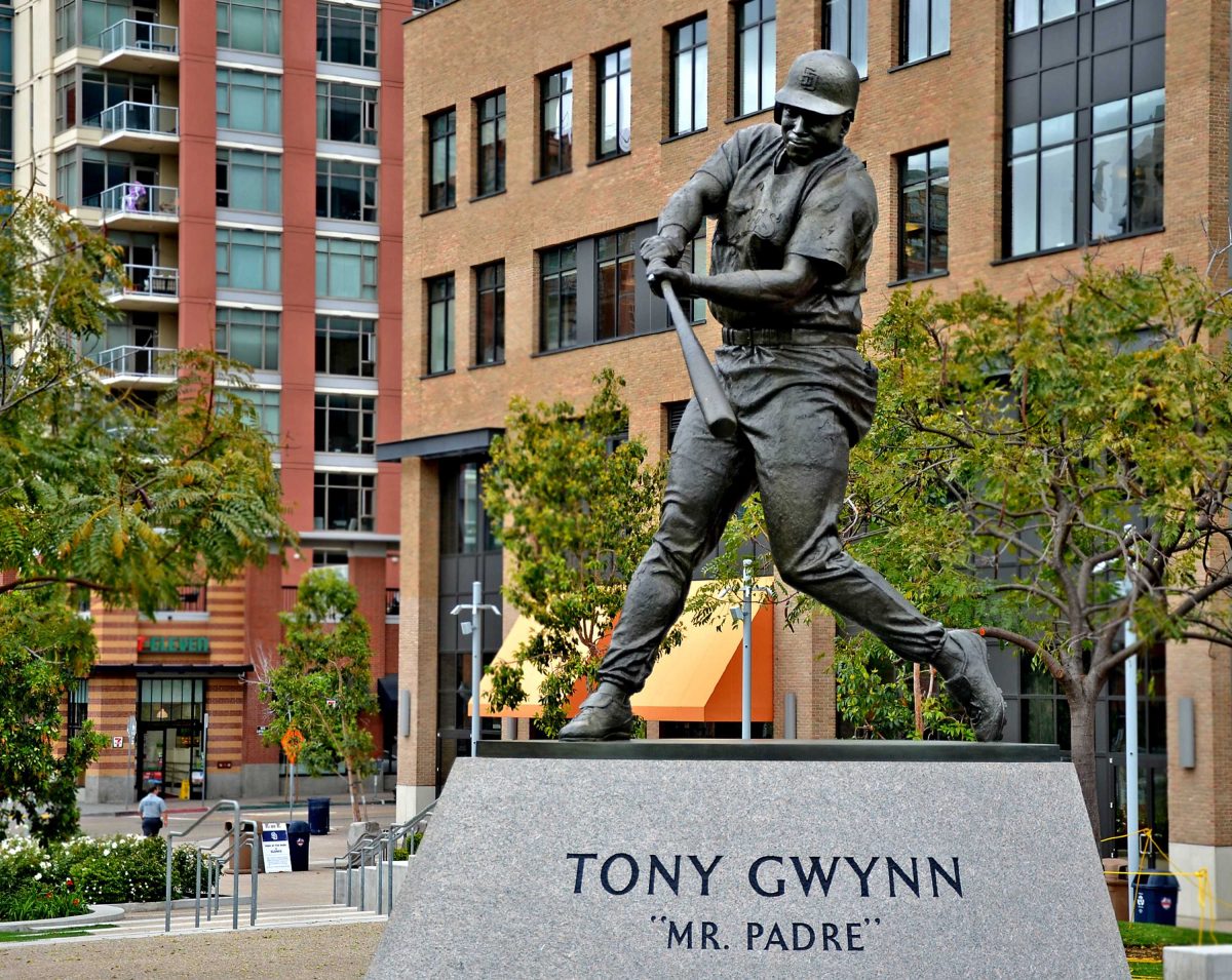 Bronze statue of San Diego Padres Tony Gwynn. He swings a baseball bat in midswing with his right foot in front of the other. An office and an apartment building are in the background in Downtown San Diego.