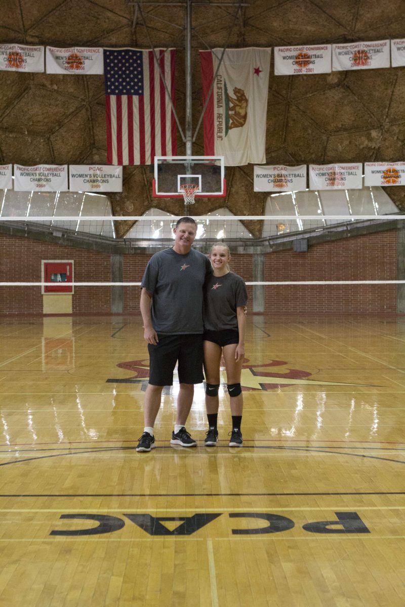 Palomar volleyball player Mikayla Seiler (right) stands next to her father, Coach Seiler, in front of a net in The Dome. They both wear gray T-shirts with a Palomar star logo and black shorts.