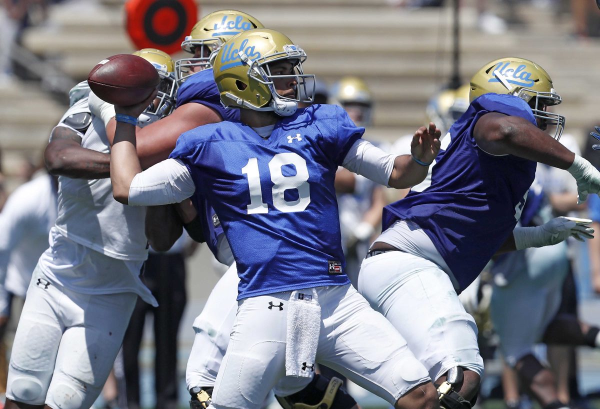 A football player brings his right hand back behind his head to throw a football as other players tacklet each other behind him.