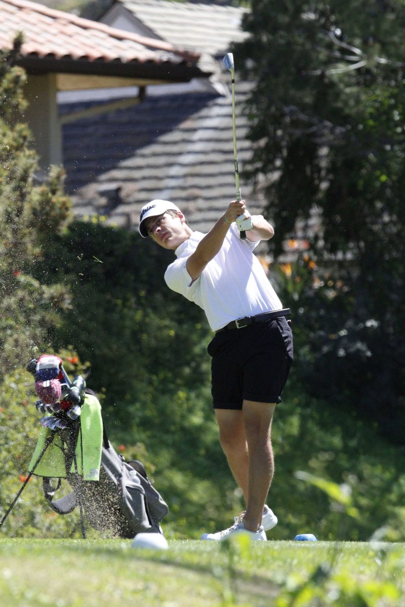 Palomar golfer Jacob Montes swings a golf club. Dirt flies to the left in front of a golf bag full of golf clubs. He wears a white polo shirt, white cap, and black shorts.