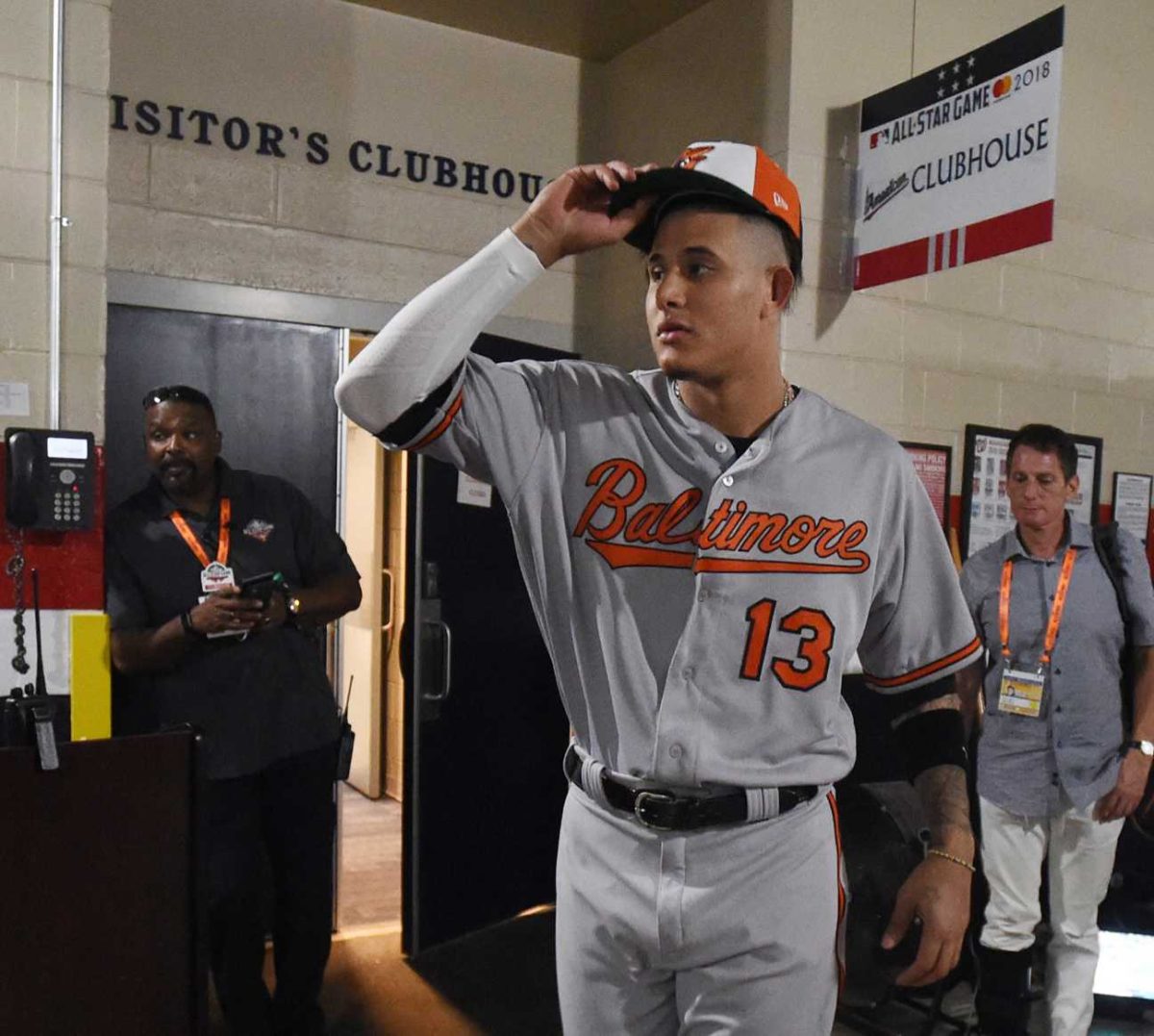 A Baltimore Oriole baseball player walks in a hallways with several people behind him. His puts on a cap with his right hand.