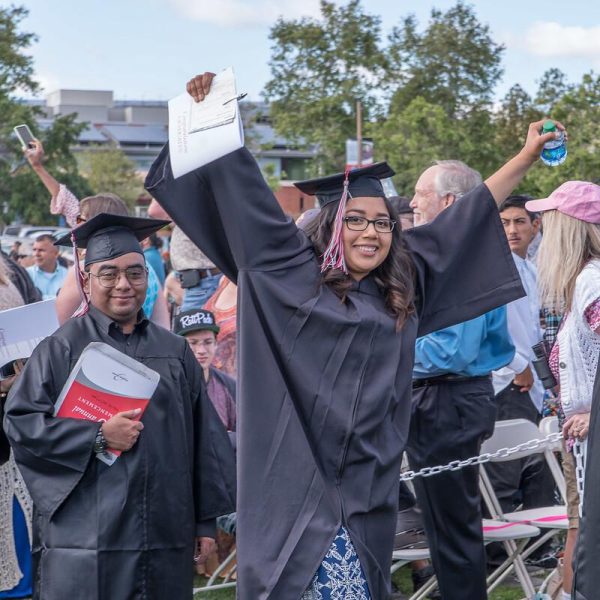 A female student in a cap and gown raises her hands in the air, holding a dipolma in her right hand and a bottle of water in her left hand.